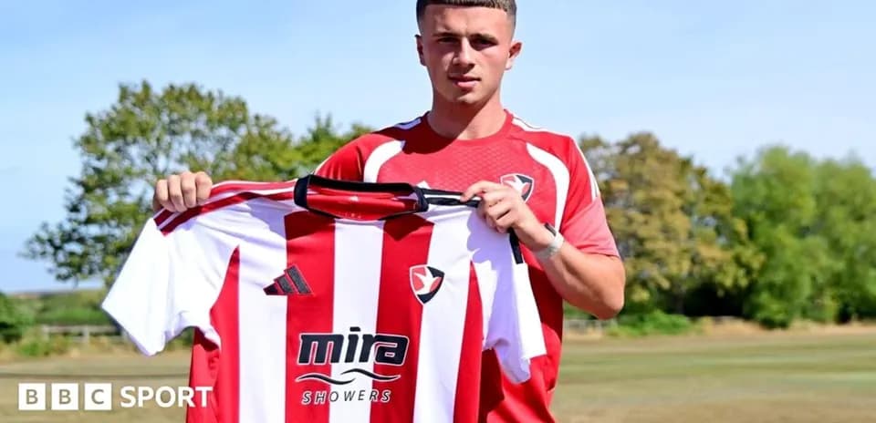 Игрок держит футболку клуба Cheltenham Town на фоне поля