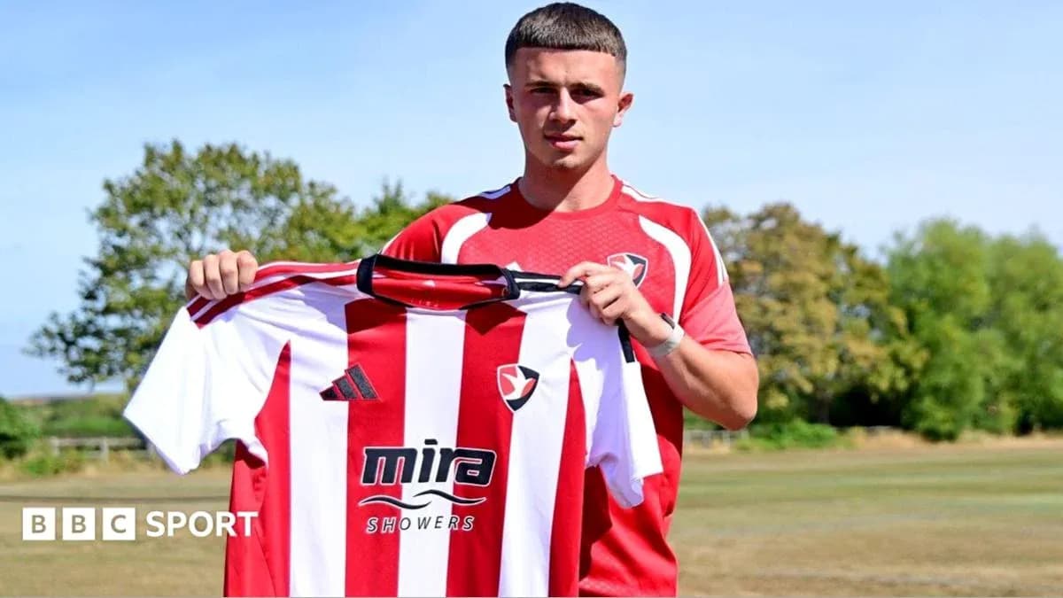 Игрок держит футболку клуба Cheltenham Town на фоне поля